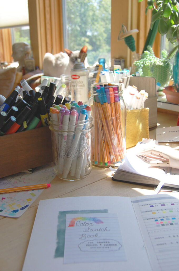 View of a sunny desk filled with various bins and jars of markers, with a hand made color swatch book open in the foreground.